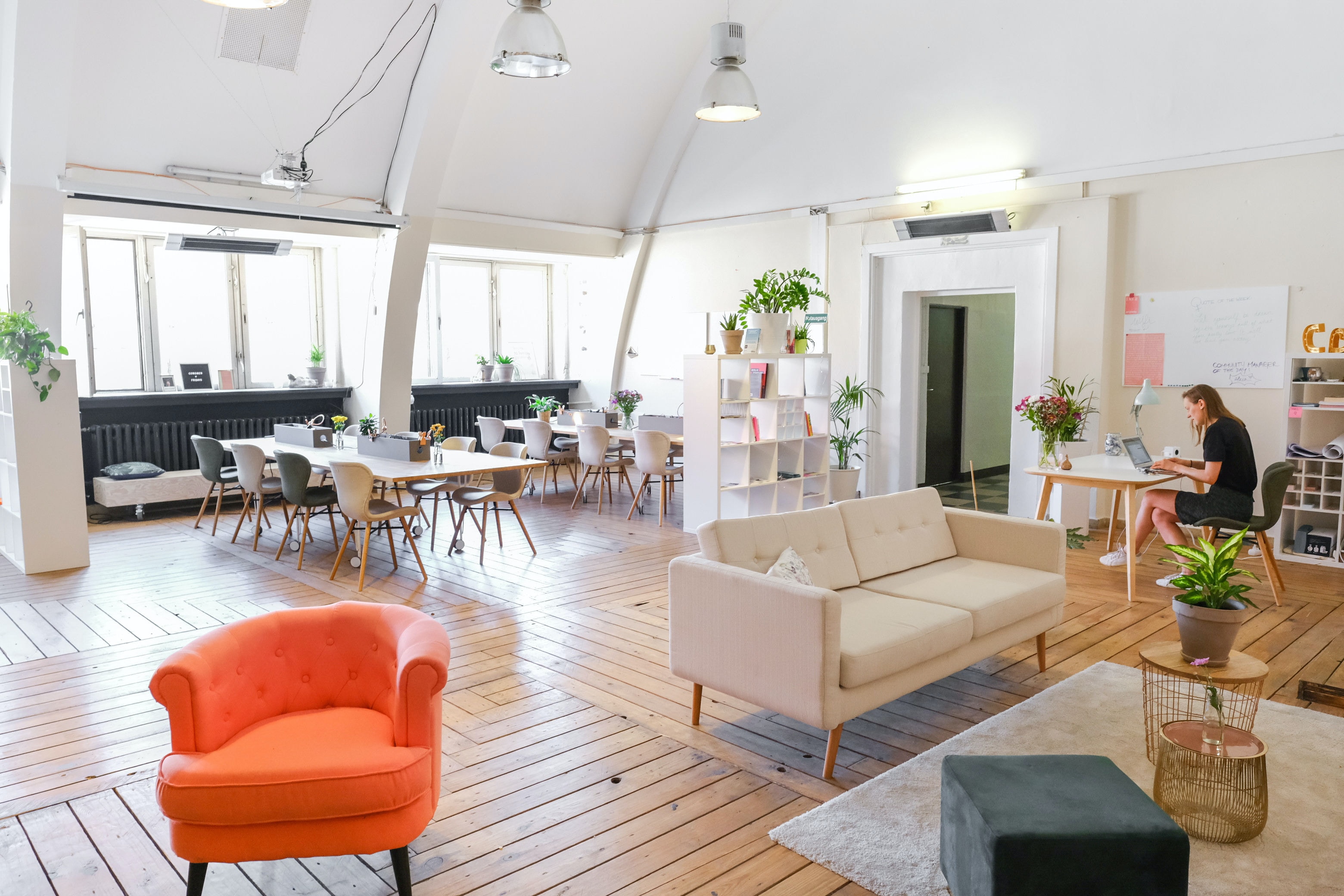 An open office space with tables and desks, comfy couches, and decorations including plants, books and lighting.