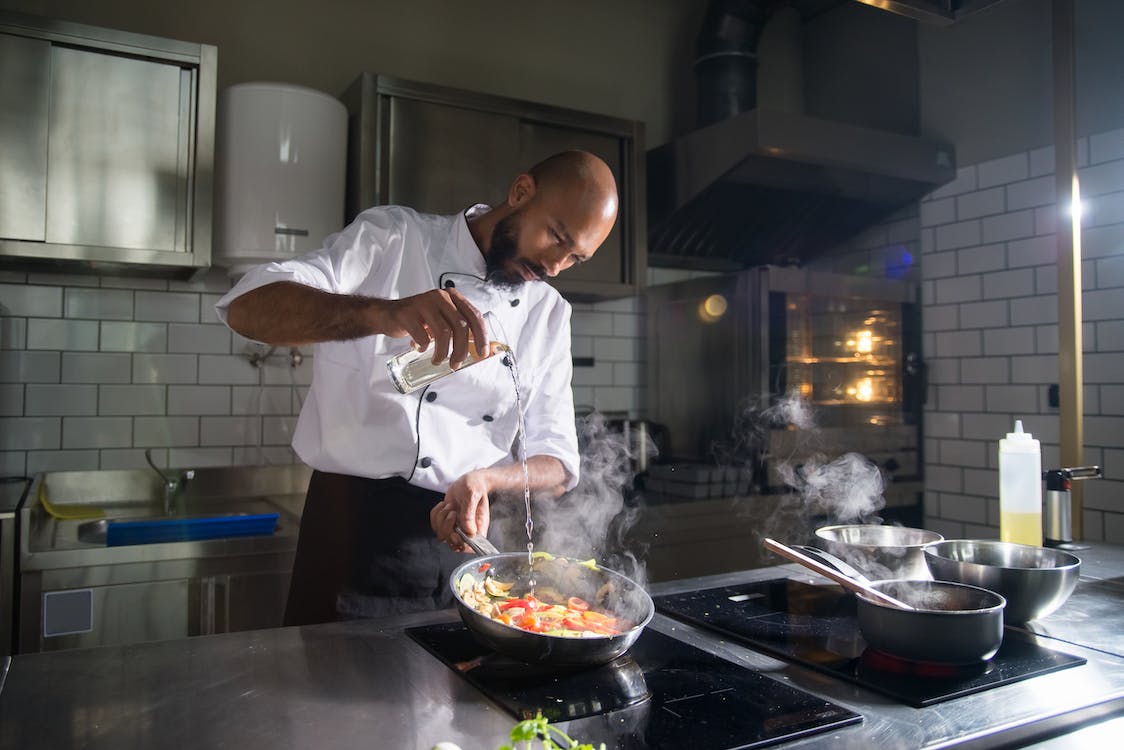 Chef cooking in a hot pan on top of a black cooker in restaurant kitchen. The surfaces are made from stainless steal and the walls are white tile.